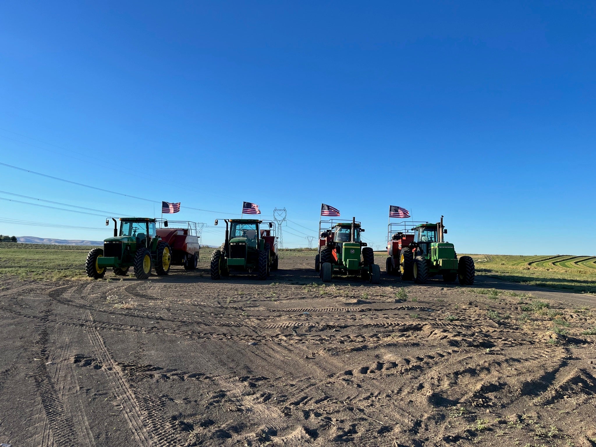 Four green tractors with American flags on a dirt field under a clear blue sky.